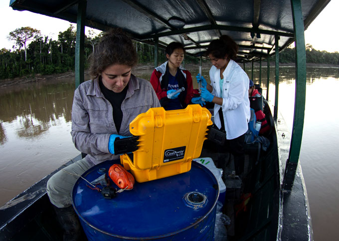 Three people are standing on a boat. Behind them are trees along the shore of the river. The person on the foreground is looking into a yellow case, atop a blue steel barrel. They are all wearing blue gloves.